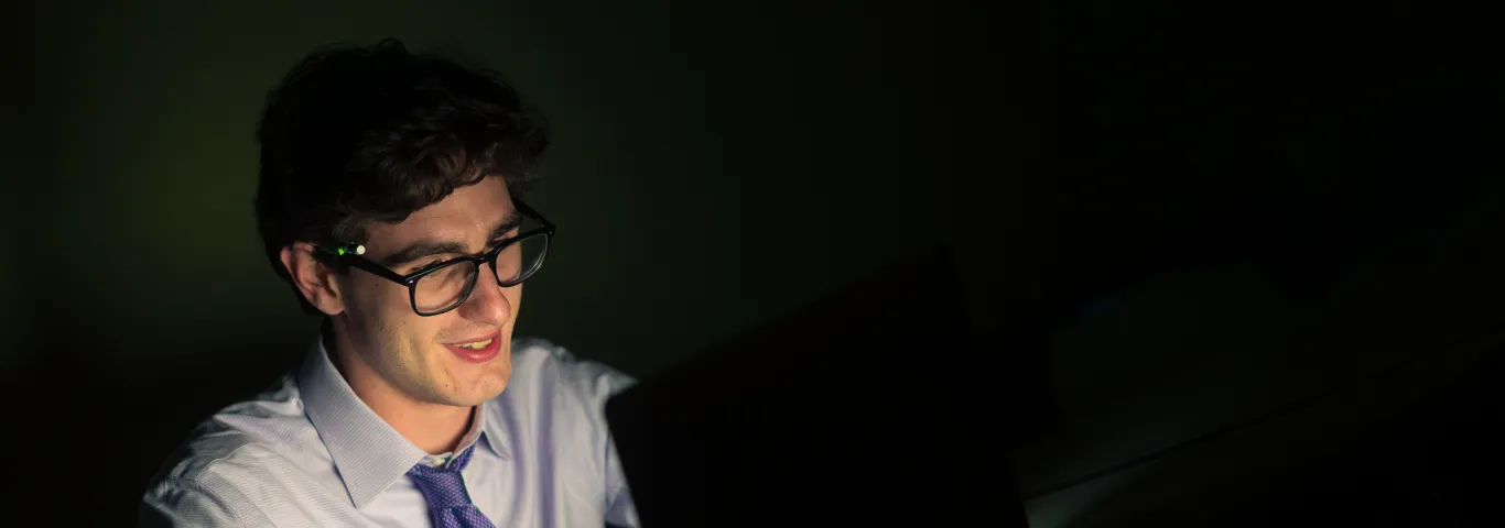 A white man with dark hair and a blue tie reads from a laptop.