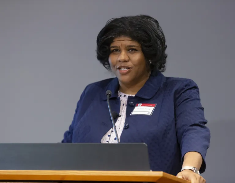 Michelle Purdy, a Black woman with dark hair and a navy blazer, speaks at a podium at the Day of Dialogue and Action