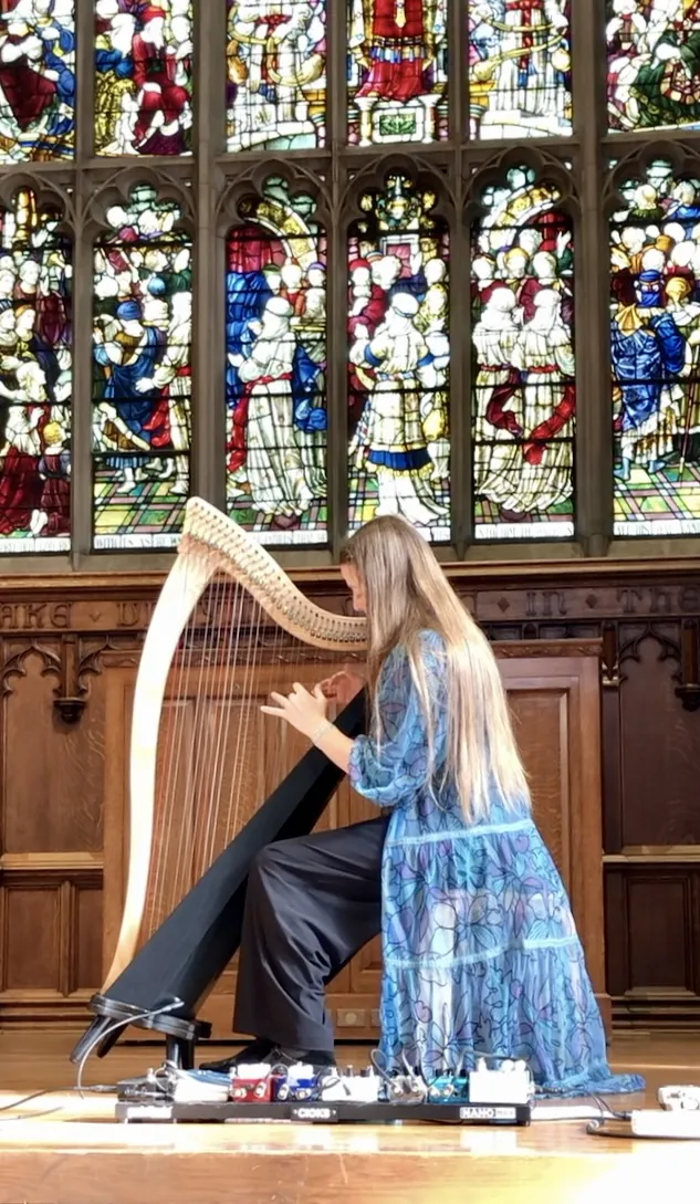 A white woman with long brown hair plays the harp in front of stained glass windows.