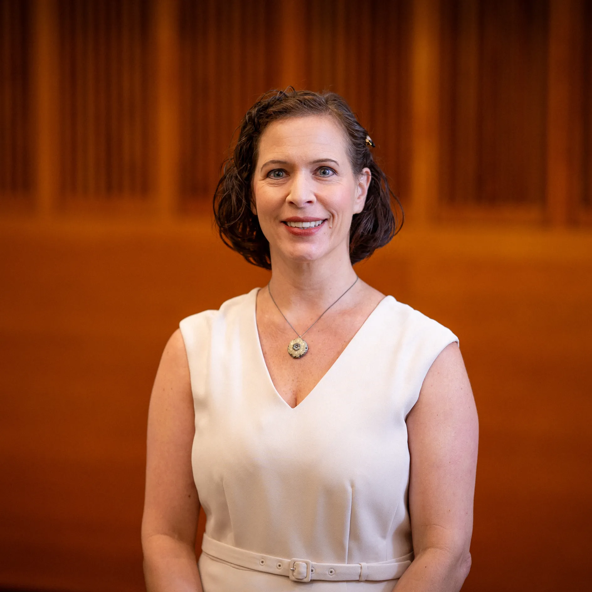 A formal headshot of Dr. Kerri Fair with wood panels in the background and a heavy vignette effect. Dr. Fair is pale with short, dark hair and wears a white dress.